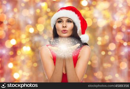 people, holidays, christmas and celebration concept - beautiful sexy woman in santa hat and red dress blowing fairy dust or snowflakes off her palms over lights background