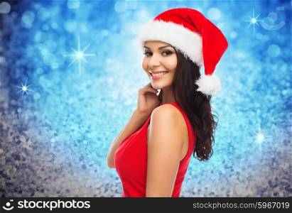 people, holidays, christmas and celebration concept - beautiful sexy woman in santa hat and red dress over blue glitter or lights background