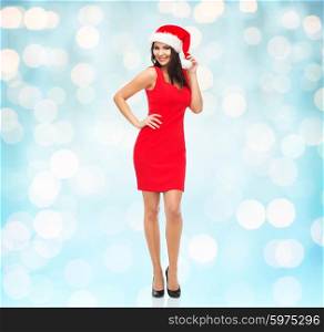 people, holidays, christmas and celebration concept - beautiful sexy woman in santa hat and red dress posing full-length over blue lights background