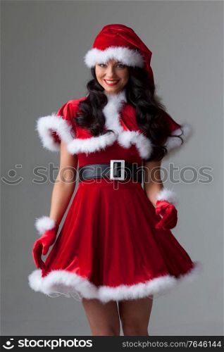 People, holidays, christmas and celebration concept - beautiful sexy woman in santa hat and red dress on gray background. Woman in santa hat and dress