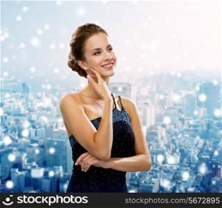 people, holidays, christmas and glamour concept - smiling woman in evening dress showing earrings over snowy city background
