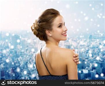 people, holidays, christmas and glamour concept - smiling woman in evening dress showing earrings over snowy city background