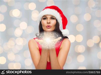 people, holidays, christmas and magic concept - beautiful sexy woman in santa hat and red dress blowing fairy dust off her palms over holidays lights background
