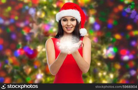 people, holidays, christmas and magic concept - beautiful sexy woman in santa hat and red dress showing fairy dust on palms over party lights background