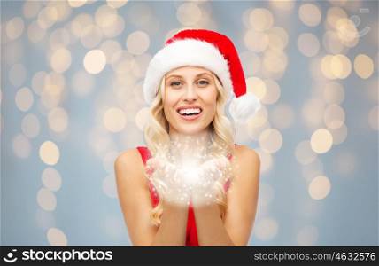 people, holidays, christmas and magic concept - happy blonde woman in santa hat holding fairy dust in palms over lights background