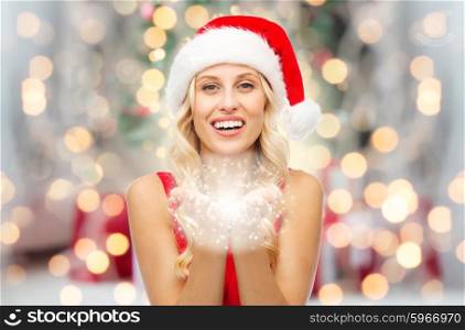 people, holidays, christmas and magic concept - happy blonde woman in santa hat holding fairy dust on palms over lights background