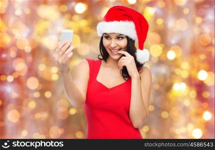 people, holidays, christmas and technology concept - beautiful sexy woman in red santa hat taking selfie picture by smartphone over lights background