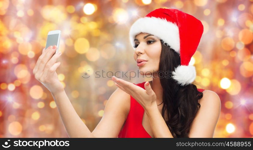 people, holidays, christmas and technology concept - beautiful sexy woman in red santa hat taking selfie picture by smartphone and sending blow kiss to camera over lights background