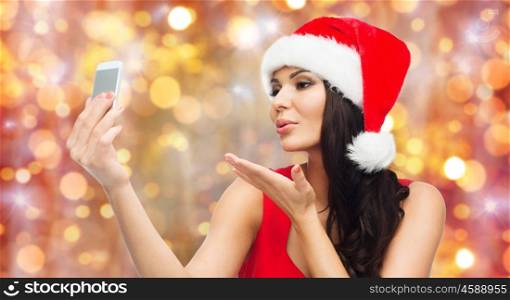 people, holidays, christmas and technology concept - beautiful sexy woman in red santa hat taking selfie picture by smartphone and sending blow kiss to camera over lights background