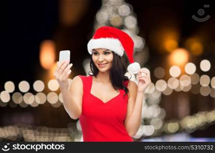 people, holidays, christmas and technology concept - beautiful sexy woman in red santa hat taking selfie picture by smartphone. woman in santa hat taking selfie by smartphone