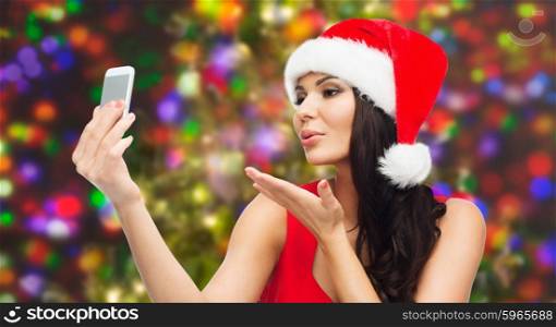 people, holidays, christmas and technology concept - beautiful sexy woman in red santa hat taking selfie picture by smartphone and sending blow kiss to camera