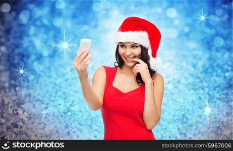people, holidays, christmas and technology concept - beautiful sexy woman in red santa hat taking selfie picture by smartphone over blue glitter or lights background