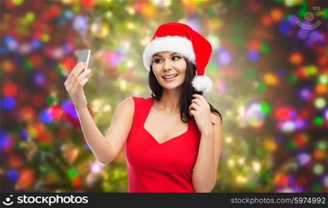 people, holidays, christmas and technology concept - beautiful sexy woman in red santa hat taking selfie picture by smartphone over party lights background