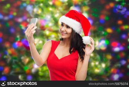 people, holidays, christmas and technology concept - beautiful sexy woman in red santa hat taking selfie picture by smartphone over party lights background