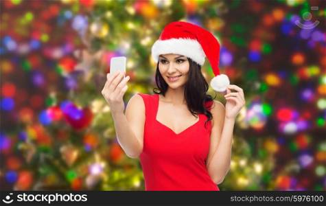 people, holidays, christmas and technology concept - beautiful sexy woman in red santa hat taking selfie picture by smartphone over holidays lights background