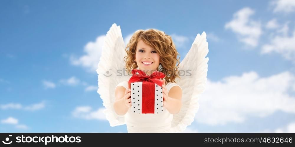 people, holidays, christmas, birthday and religious concept - happy young woman with angel wings holding gift box over blue sky and clouds background
