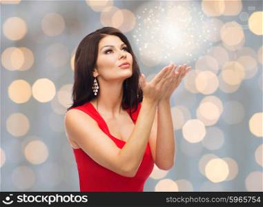 people, holidays, christmas, magic and fashion concept - beautiful sexy woman in red dress blowing fairy dust off over lights background