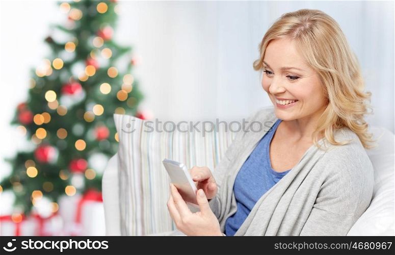 people, holidays, communication, technology and internet concept - smiling woman with smartphone texting at home over christmas tree background
