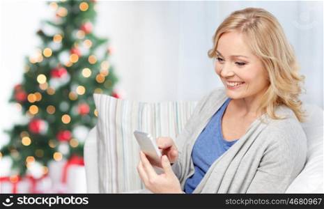 people, holidays, communication, technology and internet concept - smiling woman with smartphone texting at home over christmas tree background