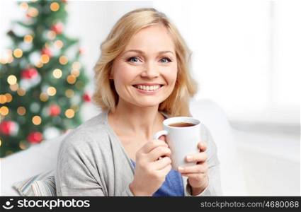 people, holidays, cosiness, drinks and leisure concept - smiling woman with cup of tea or coffee at home over christmas tree background