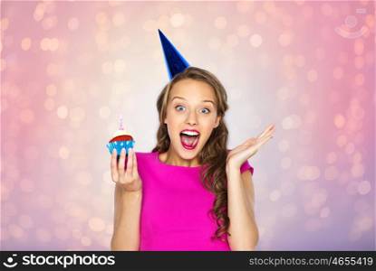 people, holidays, emotion, expression and celebration concept - happy young woman or teen girl in pink dress and party cap with birthday cupcake over rose quartz and serenity lights background