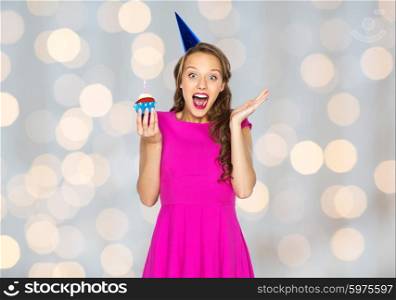 people, holidays, emotion, expression and celebration concept - happy young woman or teen girl in pink dress and party cap with birthday cupcake over lights background