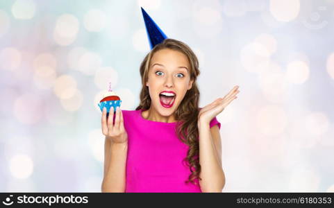 people, holidays, emotion, expression and celebration concept - happy young woman or teen girl in pink dress and party cap with birthday cupcake over holidays lights background