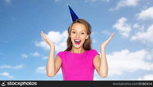 people, holidays, emotion, expression and celebration concept - happy young woman or teen girl in pink dress and party cap over blue sky and clouds background