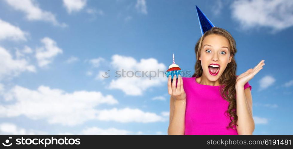 people, holidays, emotion, expression and celebration concept - happy young woman or teen girl in pink dress and party cap with birthday cupcake over blue sky and clouds background