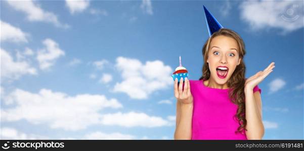 people, holidays, emotion, expression and celebration concept - happy young woman or teen girl in pink dress and party cap with birthday cupcake over blue sky and clouds background