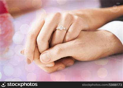 people, holidays, engagement and love concept - close up of engaged couple holding hands with diamond ring over holidays lights background