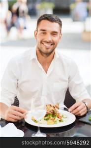 people, holidays, food and leisure concept - happy man with fork and knife eating salad for dinner at restaurant terrace