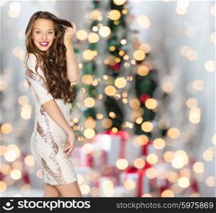 people, holidays, hairstyle and fashion concept - happy young woman or teen girl in fancy dress with sequins touching long wavy hair over christmas tree lights background