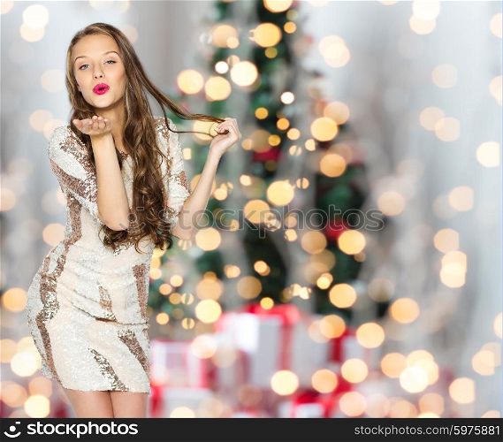 people, holidays, hairstyle and fashion concept - happy young woman or teen girl in fancy dress with sequins and long wavy hair sending blow kiss over christmas tree lights background