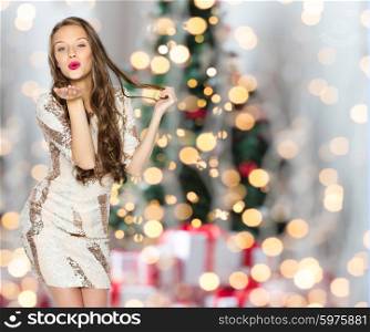 people, holidays, hairstyle and fashion concept - happy young woman or teen girl in fancy dress with sequins and long wavy hair sending blow kiss over christmas tree lights background
