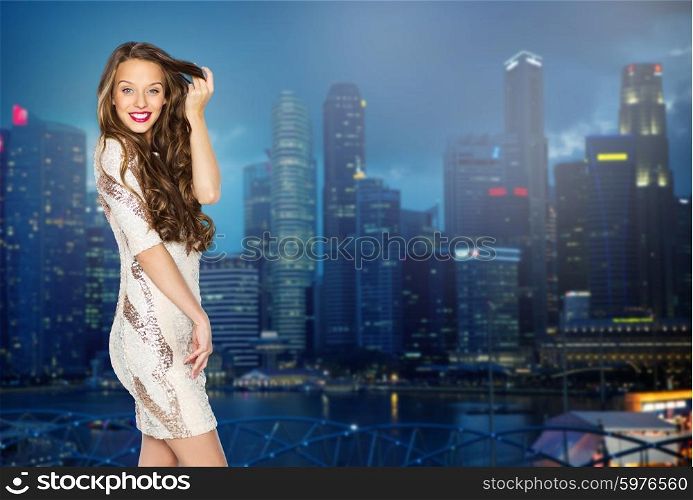 people, holidays, hairstyle, nightlife and fashion concept - happy young woman or teen girl in fancy dress with sequins touching long wavy hair over night singapore city background