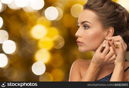 people, holidays, jewelry and luxury concept - close up of woman in evening dress fastening diamond earring over lights background
