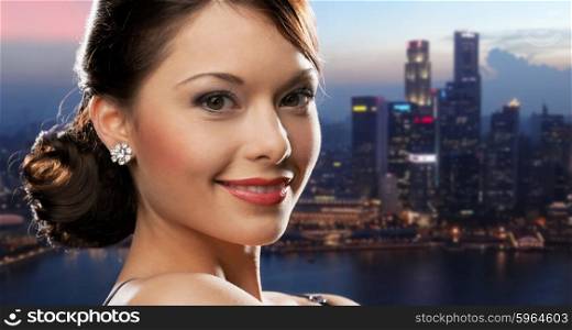 people, holidays, jewelry and luxury concept - smiling woman face with diamond earring over night singapore city background