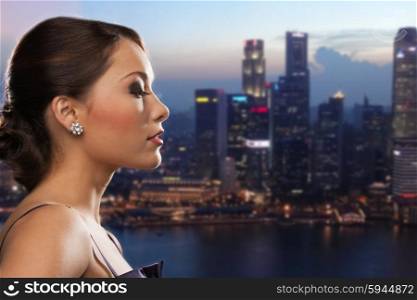people, holidays, jewelry and luxury concept - woman face with diamond earring over night singapore city background