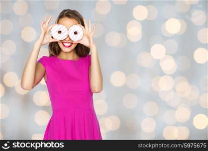 people, holidays, junk food and fast food concept - happy young woman or teen girl in pink dress having fun and looking through donuts over lights background