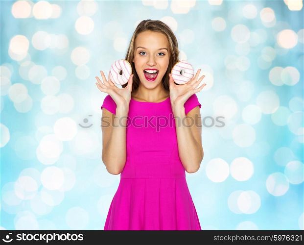 people, holidays, junk food and fast food concept - happy young woman or teen girl in pink dress with donuts over blue holidays lights background