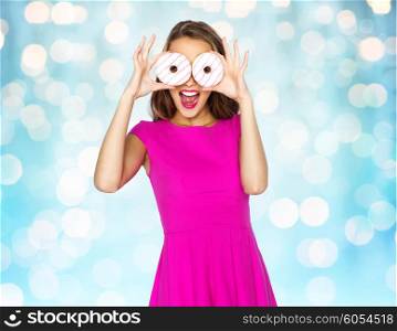 people, holidays, junk food and fast food concept - happy young woman or teen girl in pink dress having fun and looking through donuts over blue holidays lights background