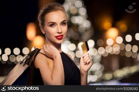 people, holidays, luxury and sale concept - beautiful woman with credit card and shopping bags over christmas tree lights background. woman with credit card and shopping bags