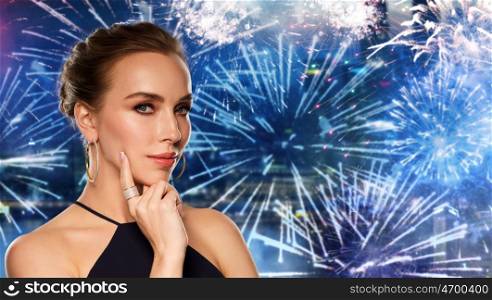 people, holidays, luxury, jewelry and fashion concept - beautiful woman in black wearing golden earrings and ring with diamonds over night city firework background