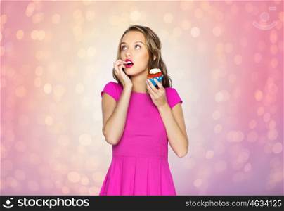 people, holidays, party, junk food and celebration concept - happy young woman or teen girl in pink dress eating birthday cupcake over rose quartz and serenity lights background