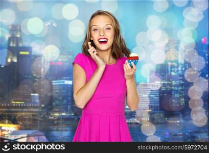 people, holidays, party, junk food and celebration concept - happy young woman or teen girl in pink dress with birthday cupcake over night city and holidays lights background