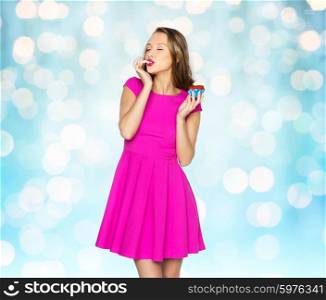 people, holidays, party, junk food and celebration concept - happy young woman in pink dress eating birthday cupcake over blue holidays lights background