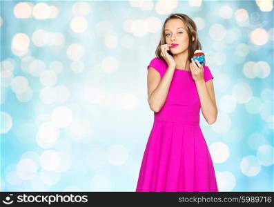people, holidays, party, junk food and celebration concept - happy young woman or teen girl in pink dress with birthday cupcake over blue holidays lights background