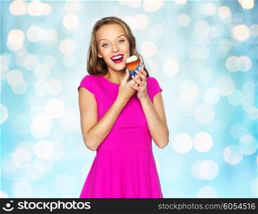 people, holidays, party, junk food and celebration concept - happy young woman or teen girl in pink dress with cupcake over blue holidays lights background