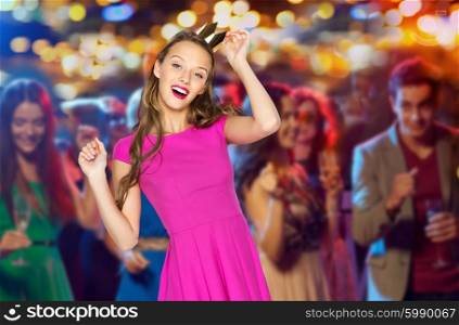people, holidays, party, night life and fashion concept - happy young woman or teen girl in pink dress and princess crown dancing at disco club over crowd lights background